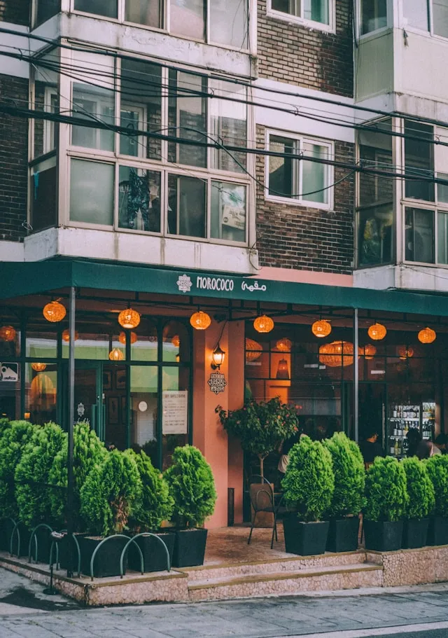 モロッコ料理レストラン「MOROCCO」の店舗正面。オレンジ色のエントランスドア、テラコッタ色のファサード。複数のオレンジ色の提灯ランタンが天井に装飾。前面に樹木のような緑色のトピアリーが大きなプランターに植えられている。上階はレンガ造りと白いバルコニー、複数の窓。アラビア文字と英文サインが表示されたモダンな建築